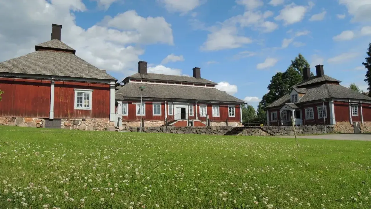 Malingsbo herrgård med sina röda trähus och stenfundament, omgivet av en grönskande äng med vita blommor under en blå himmel med moln.