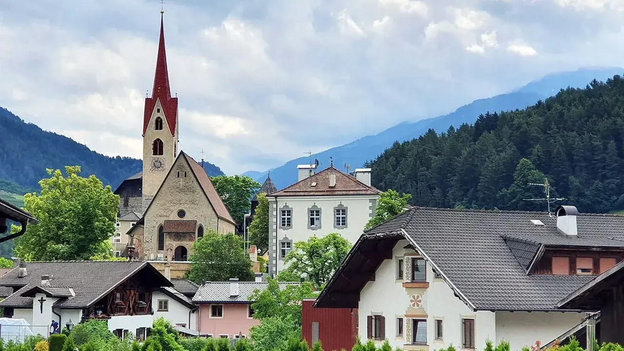 Un borgo pittoresco in Val d'Isarco, con una chiesa dal campanile rosso svettante tra case tradizionali e montagne boscose.
