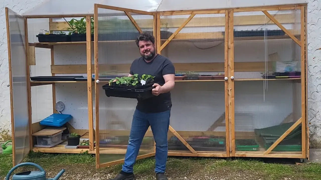 Un homme souriant tient un plateau de jeunes plants devant sa serre pour semis fait maison, remplie d'étagères de semis.