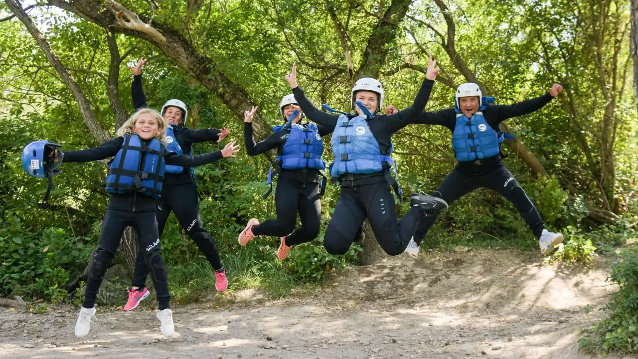 Cinco personas, incluyendo niños, saltan alegres con trajes de neopreno y chalecos salvavidas en un entorno natural, listos para una aventura en Huesca.