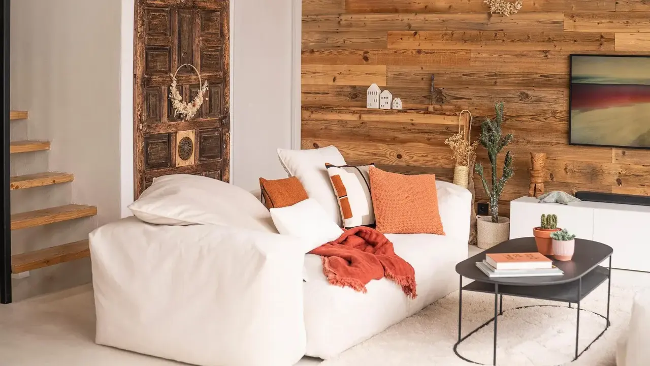 Un salon chaleureux et accueillant avec un grand canapé blanc, des coussins orange et un mur en bois rustique. Une porte ancienne décore le mur.