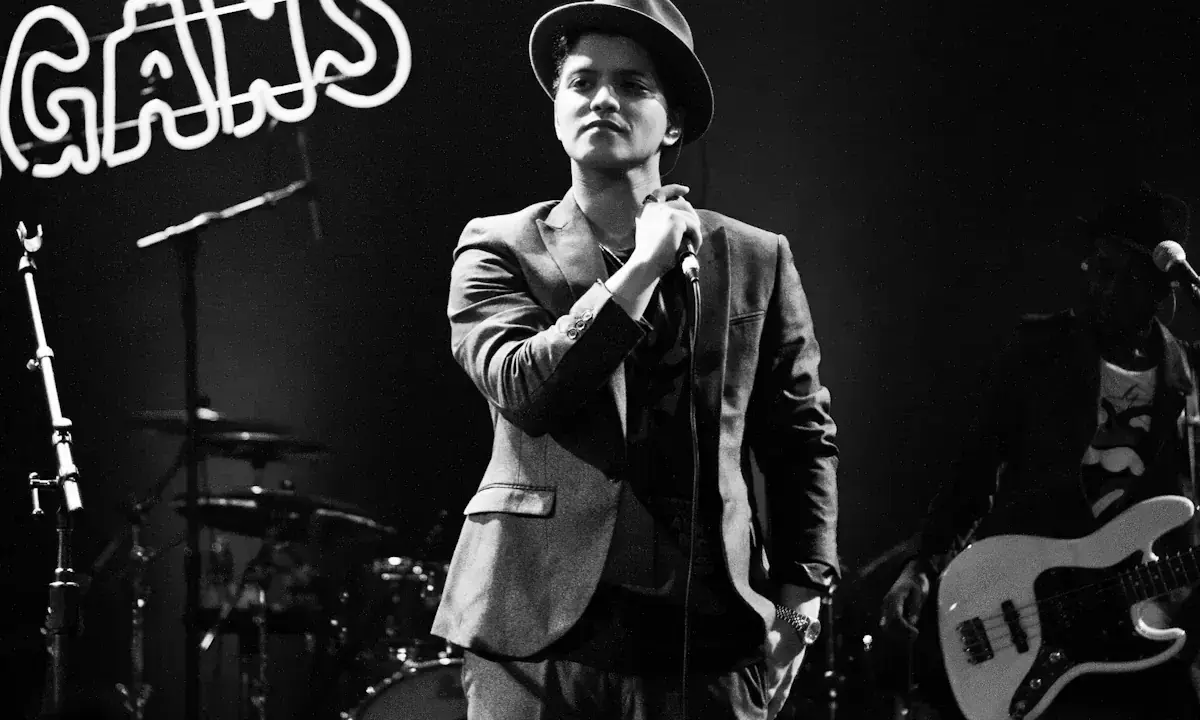 Bruno Mars i Sverige, med hatt och kavaj, sjunger i mikrofonen. Bakom honom syns ett band och neonljus.