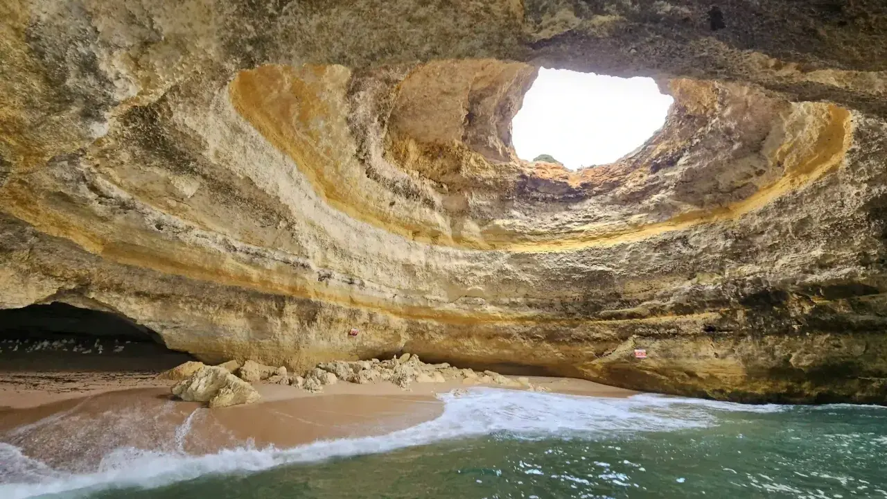 Die Benagil-Höhle mit ihrem kreisförmigen Loch im Felsdach, das Sonnenlicht hereinlässt. Wellen brechen sanft am Sandstrand im Inneren.