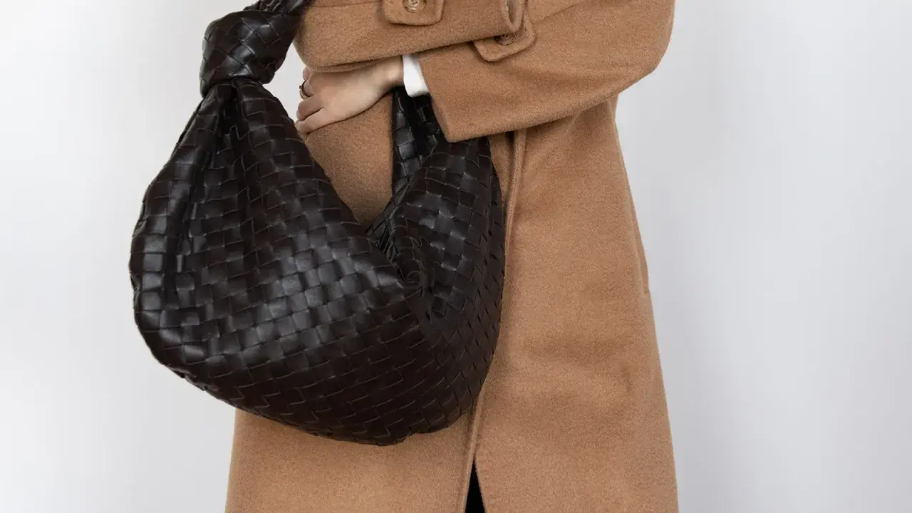 Dunkle, gewebte Tasche mit Knoten-Detail, getragen zu einem Kamelhaarmantel. Sieht aus wie eine Bottega Veneta, aber es ist ein Bottega Veneta Fake.