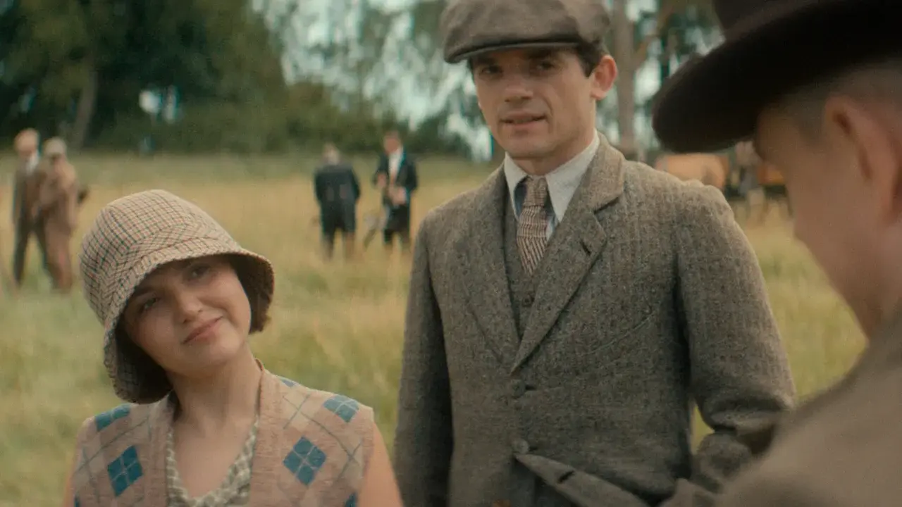 Giovane donna con cappello a scacchi e gilet a rombi, accanto a un uomo con berretto e giacca di tweed. Sullo sfondo, i sette quadranti di un campo.