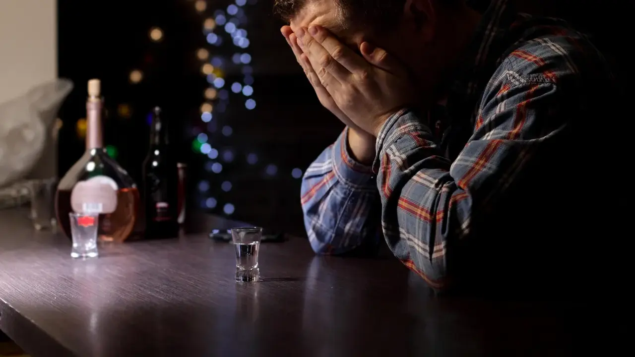 Mężczyzna z rękami na twarzy przy barze, obok butelka i kieliszek. Obraz symbolizuje przymusowe leczenie alkoholika i jego walkę.