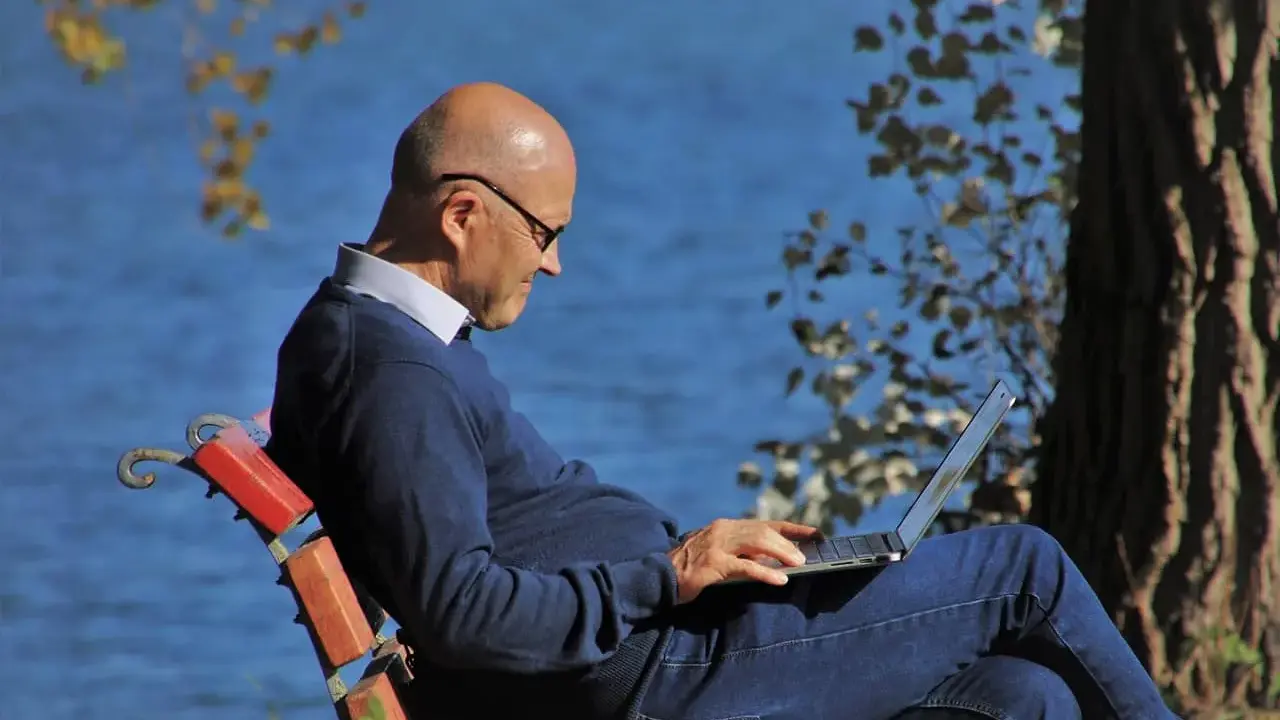 Mężczyzna w okularach siedzi na ławce nad jeziorem, pracując na laptopie. Planuje swoje ubezpieczenie emerytalne, ciesząc się spokojem natury.
