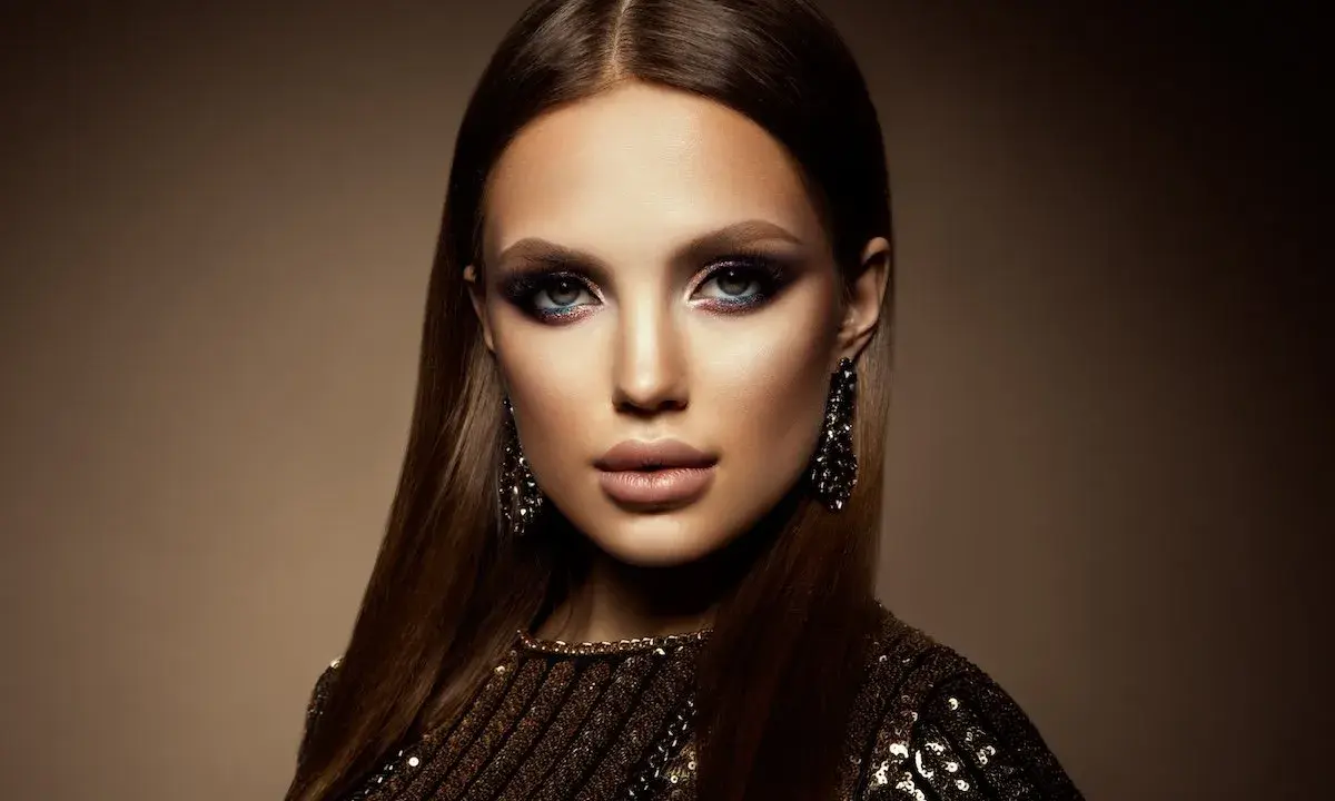 Mujer con un look de **soft goth makeup**, ojos ahumados y labios definidos, luciendo pendientes largos y un vestido brillante.