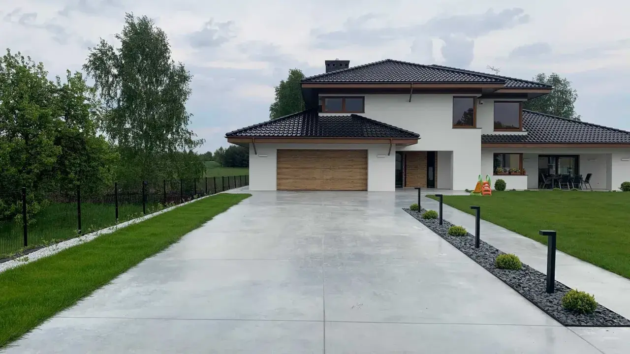 Nowoczesny dom z drewnianą bramą garażową, otoczony zielenią. Szeroki podjazd betonowy prowadzi do wejścia, a wzdłuż niego biegną lampy i rabaty z kamieniami.