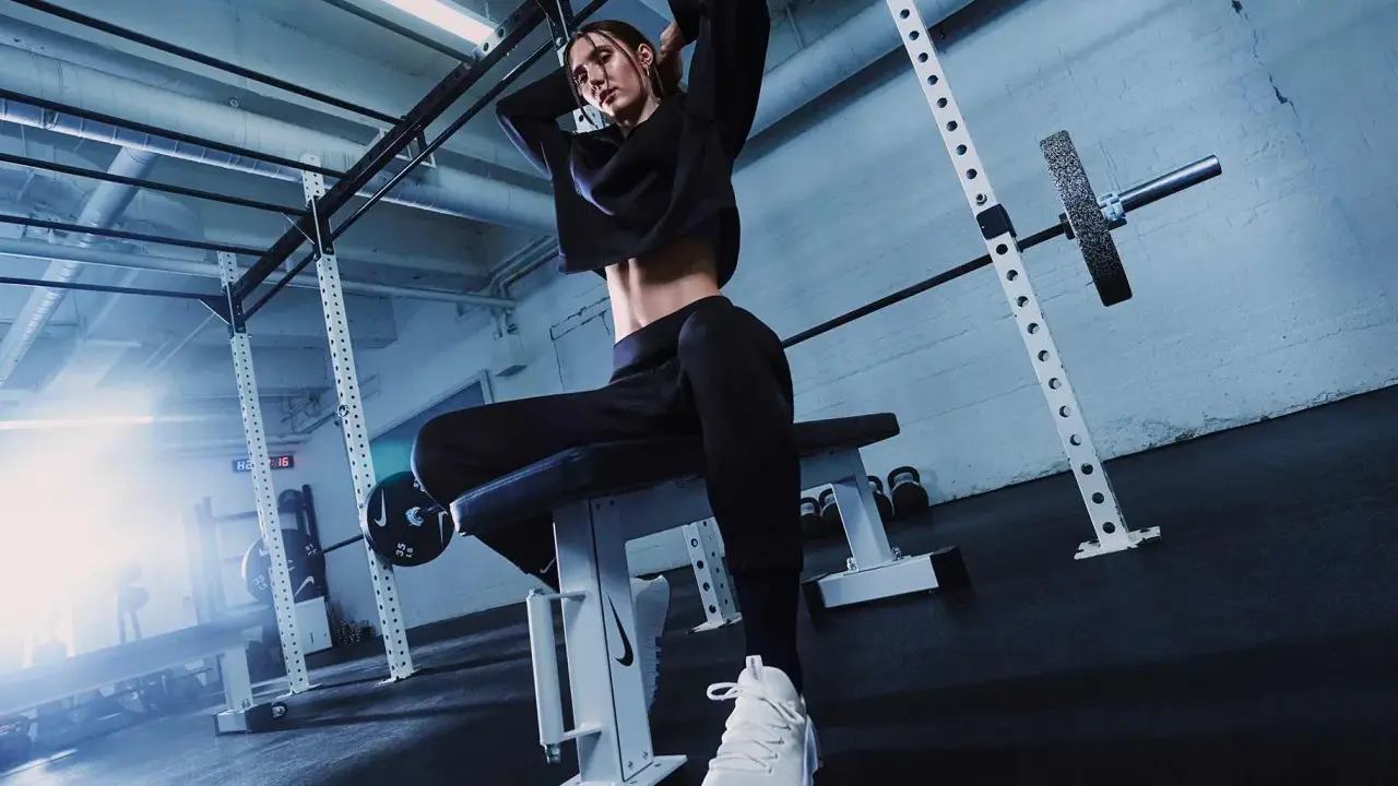 Donna in tenuta sportiva nera, con scarpe bianche, seduta su panca in palestra. Ispirazione su come vestirsi in palestra.