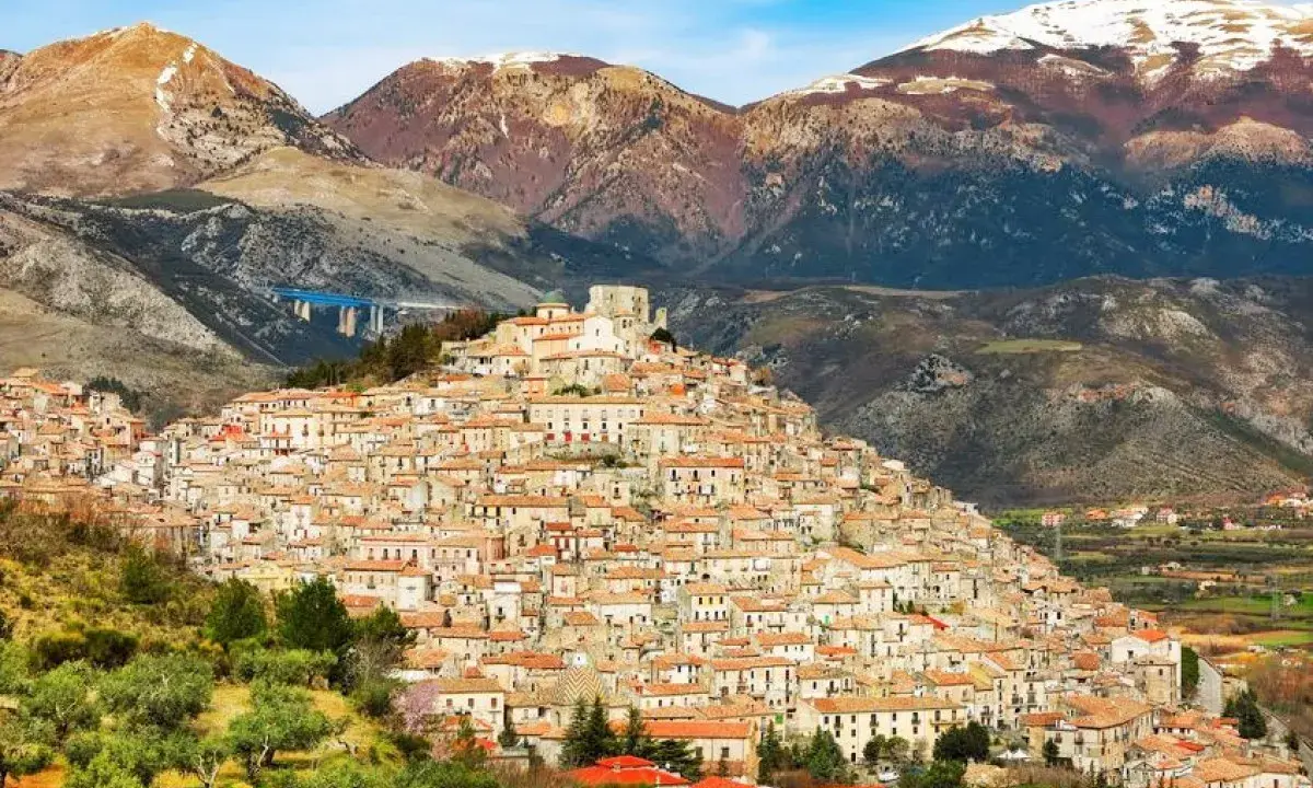 Un pittoresco borgo calabrese si aggrappa a una collina, con case in pietra e tetti rossi che si estendono fino alle montagne innevate.