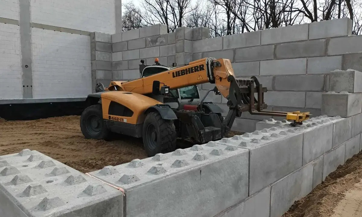 Żółty ładowacz teleskopowy Liebherr pracuje przy budowie muru oporowego z betonowych bloków.