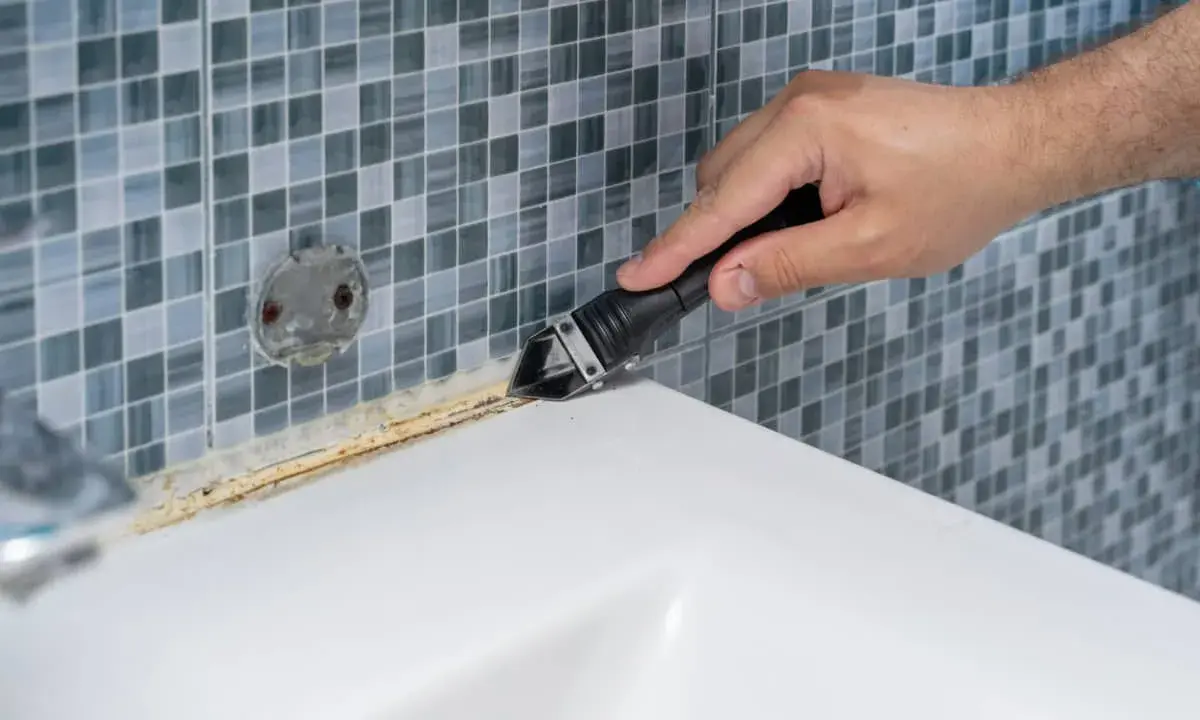 Mano usando una espátula para quitar silicona vieja y sucia de la bañera.