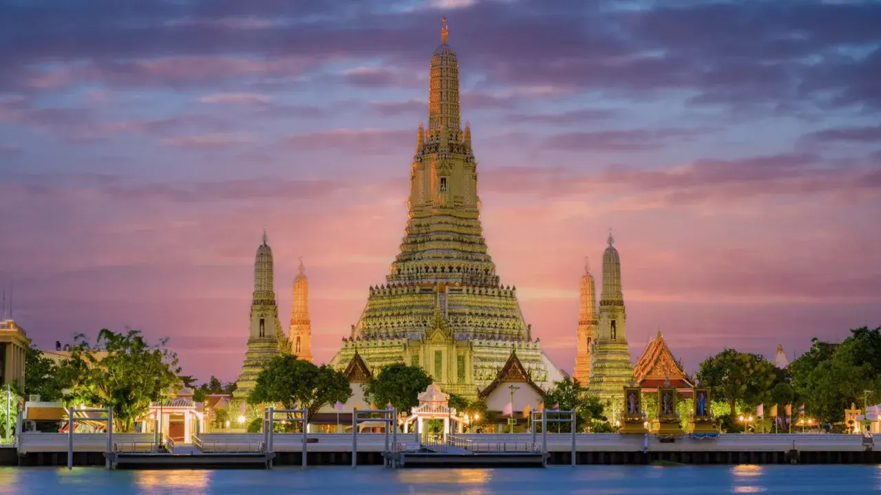 Il **tempio dell'alba Bangkok** (Wat Arun) si erge maestoso al tramonto, con le sue guglie dorate che brillano contro un cielo rosa e viola.
