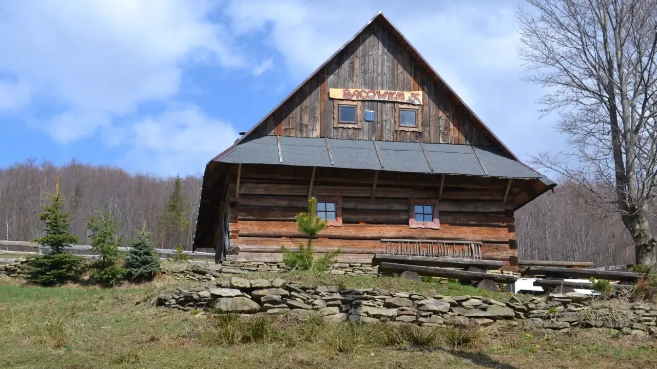Drewniana bacówka górska ostoja z tabliczką na dachu. Otoczona zielenią i kamiennym murkiem.