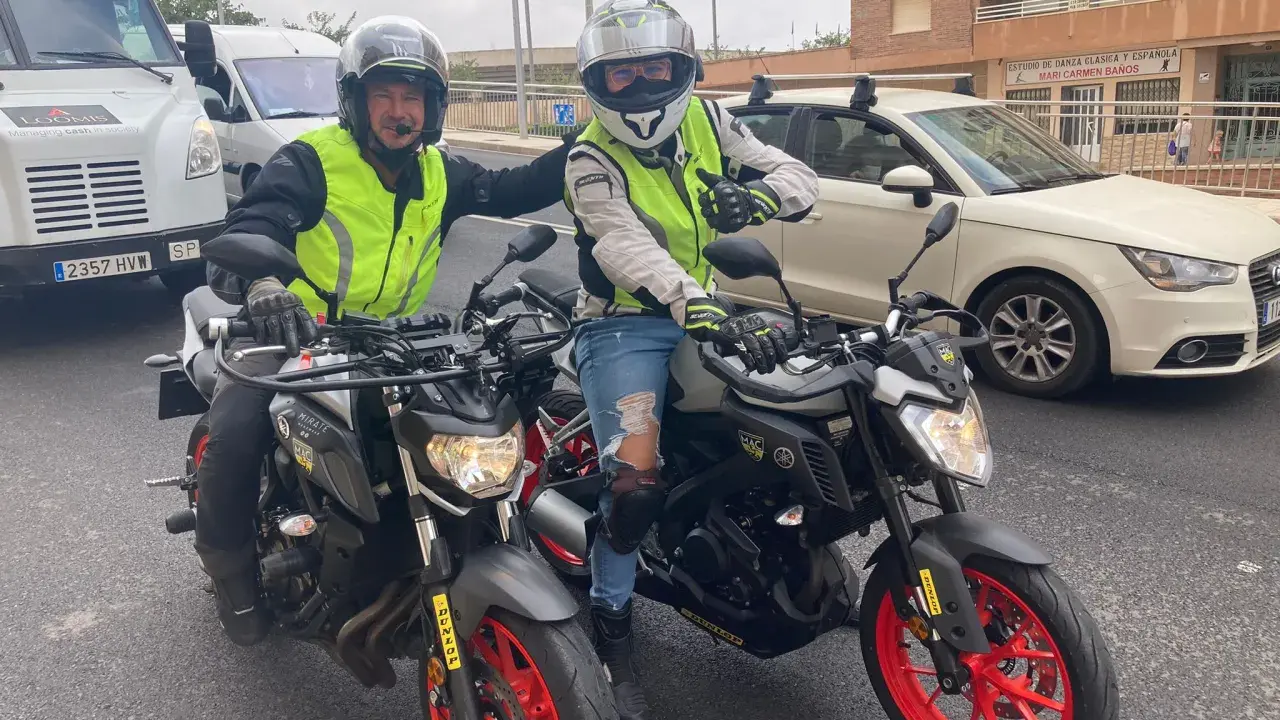 Dos motoristas con chalecos reflectantes y cascos posan con sus motos. Uno hace un gesto de "pulgar arriba". ¡Listos para la aventura en su a1 de moto!