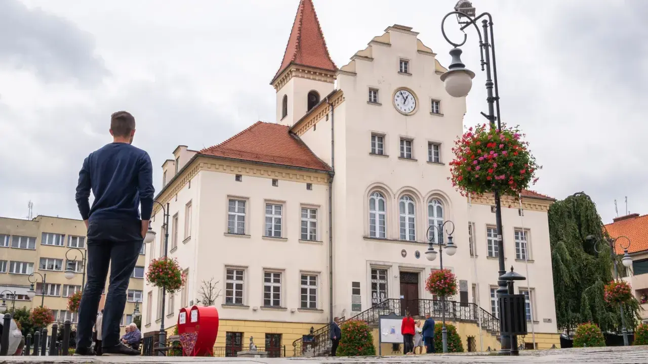 Mężczyzna podziwia ratusz, jedną z głównych trzebnickich atrakcji. W tle czerwone serce i kwitnące drzewka.