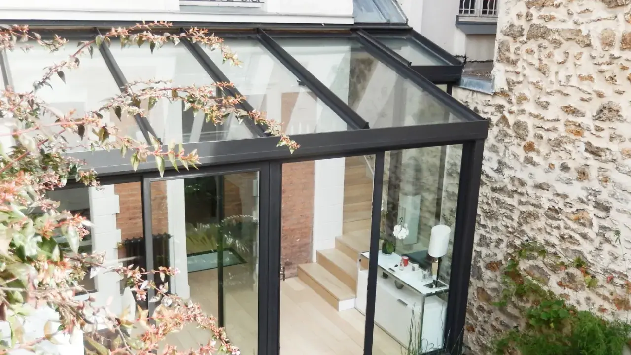 Une véranda bioclimatique moderne, avec une structure en verre et métal noir, s'intègre à une maison en briques et pierre. Avis positif sur son esthétique.