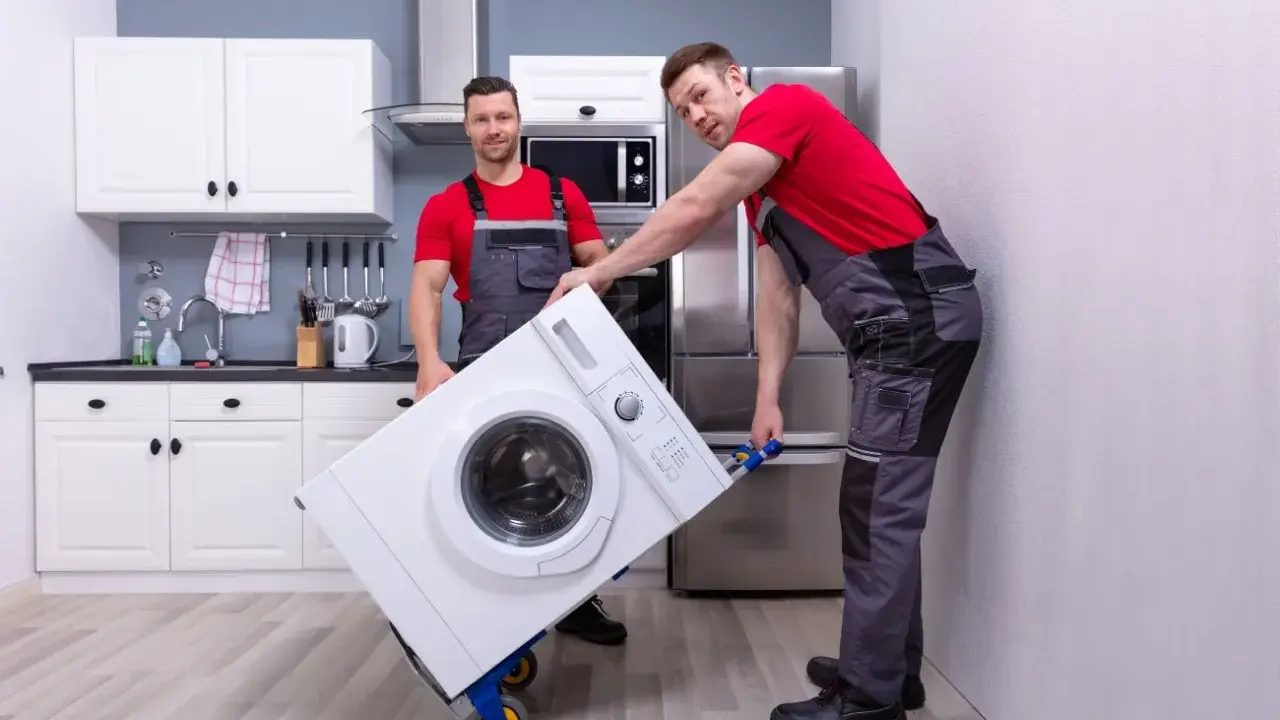 Dwóch mężczyzn w roboczych ubraniach przewozi pralkę na wózku. Czy można przewozić pralkę na leżąco? To pytanie zadaje sobie wielu przy przeprowadzce.