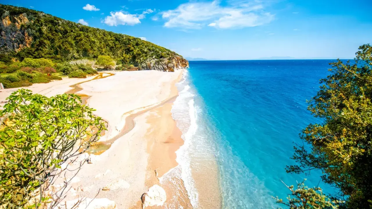En orörd albanien strand med turkost vatten, vit sand och gröna kullar. En liten flod rinner ut i havet.