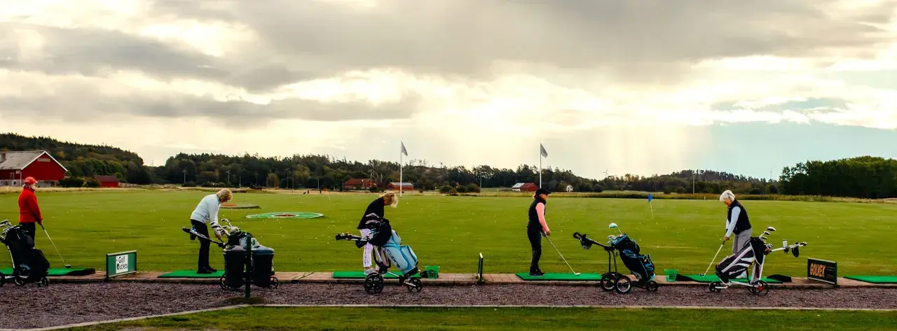 Kvinnor tränar golf på en driving range i Sverige. Solstrålar bryter igenom molnen.