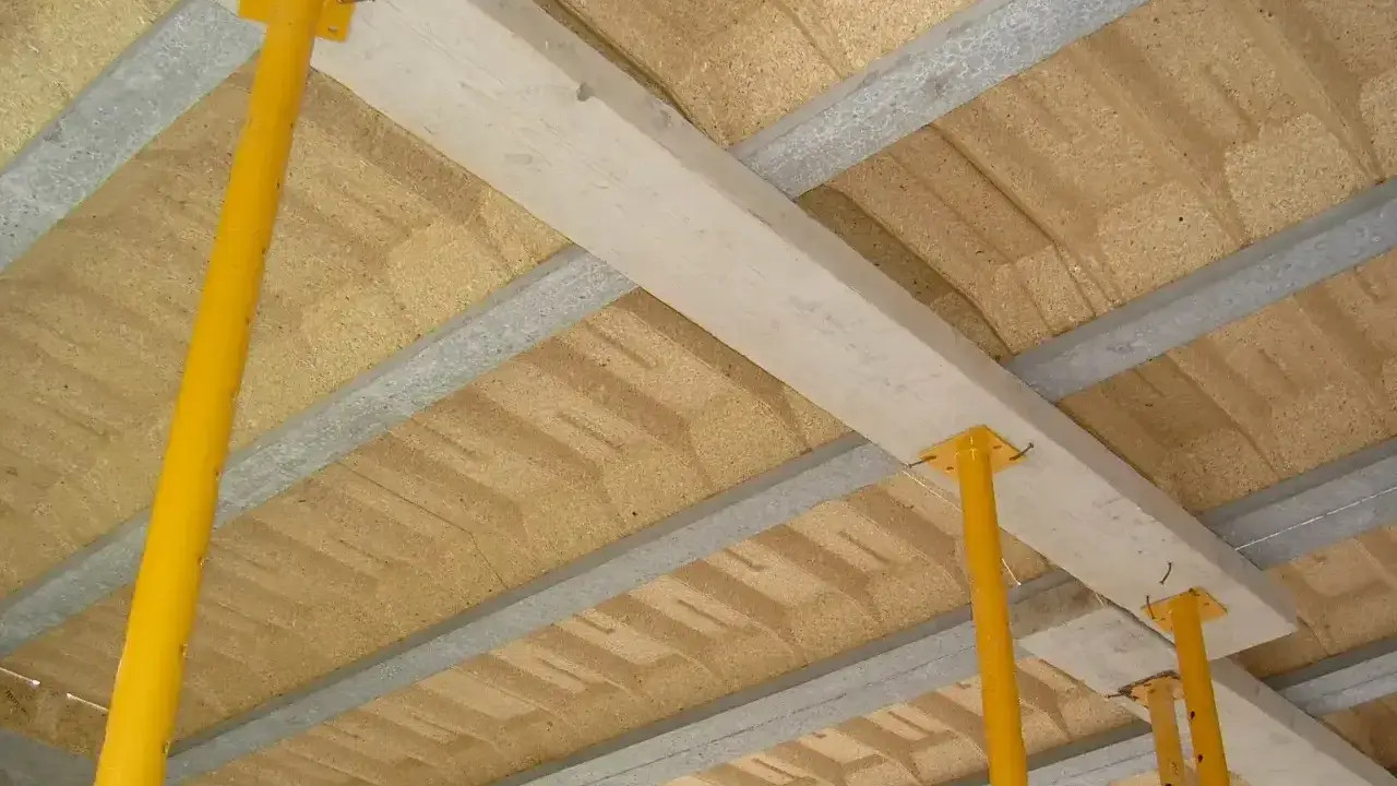 Drewniana belka stropowa podparta żółtymi stemplami, tworząca konstrukcję stropu nad płytą z materiału drewnopochodnego.