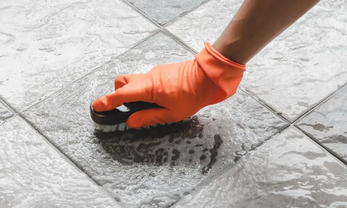 Mano con guante naranja cepillando un suelo antideslizante con espuma.