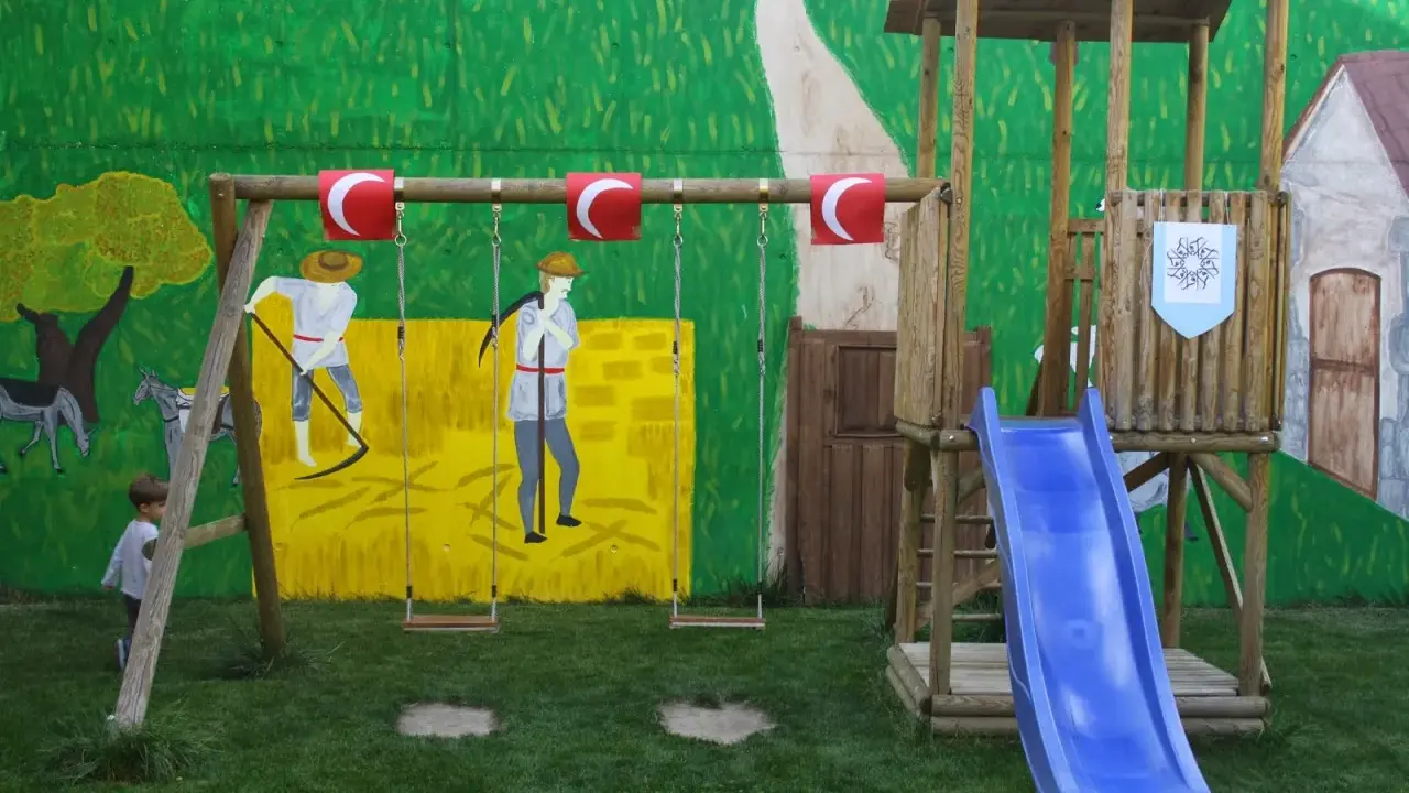 Un niño observa un columpio y un tobogán azul, parte de unos juegos medievales para niños, con un mural de fondo.