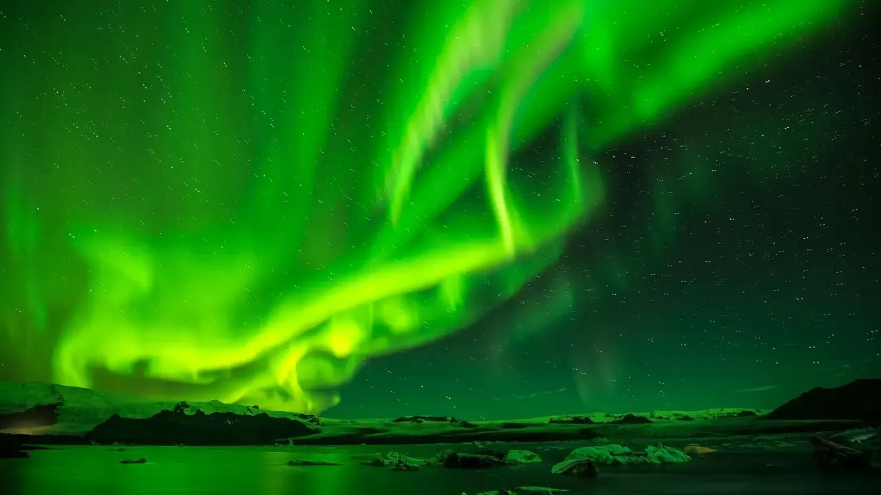 Grön norrsken dansar över en isig ö med stjärnor som glittrar. Vattnet speglar det magiska skådespelet.