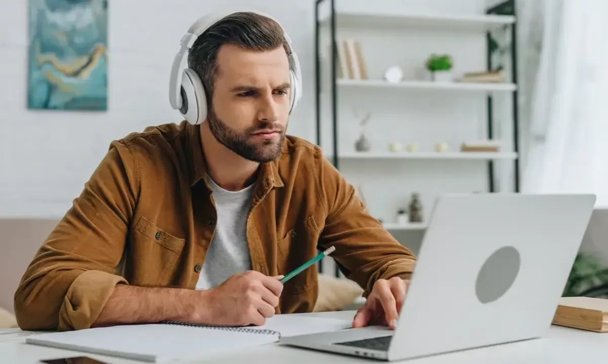 Mann mit Kopfhörern lernt online, nutzt Laptop und Notizbuch. Er wirkt konzentriert, während er mit einem Stift tippt. Ein typisches Bild für das heutige "Home-Office"-Zeitalter, das viele Anglizismen mit sich bringt.