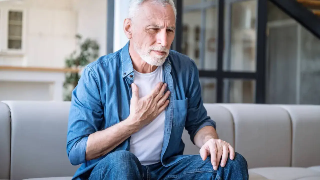 Älterer Mann mit COPD hält sich die Brust. Er sitzt auf einem Sofa und sieht besorgt aus.