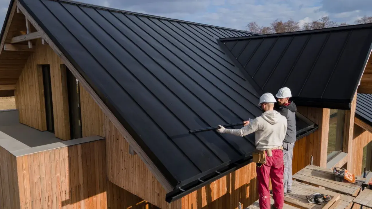 Zwei Handwerker sanieren ein Dach mit schwarzer Trapezblechdeckung. Das Holzhaus erhält ein modernes Aussehen.