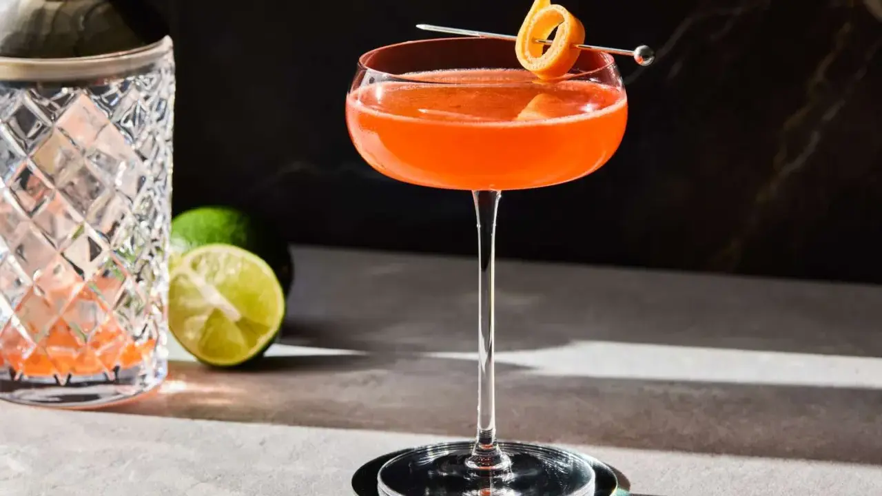 En vackert orangefärgad Division Bell cocktail med en apelsinzest som garnityr, bredvid en cocktailshaker och limeklyftor.