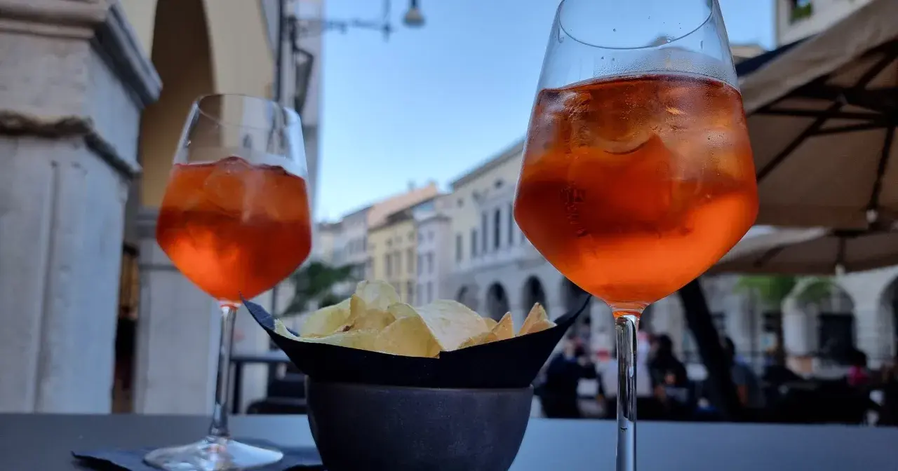 Dwa drinki Aperol Spritz i miseczka chipsów na stoliku. Czy alkohol podnosi cholesterol? To pytanie zadaje sobie wielu, delektując się takimi przekąskami.