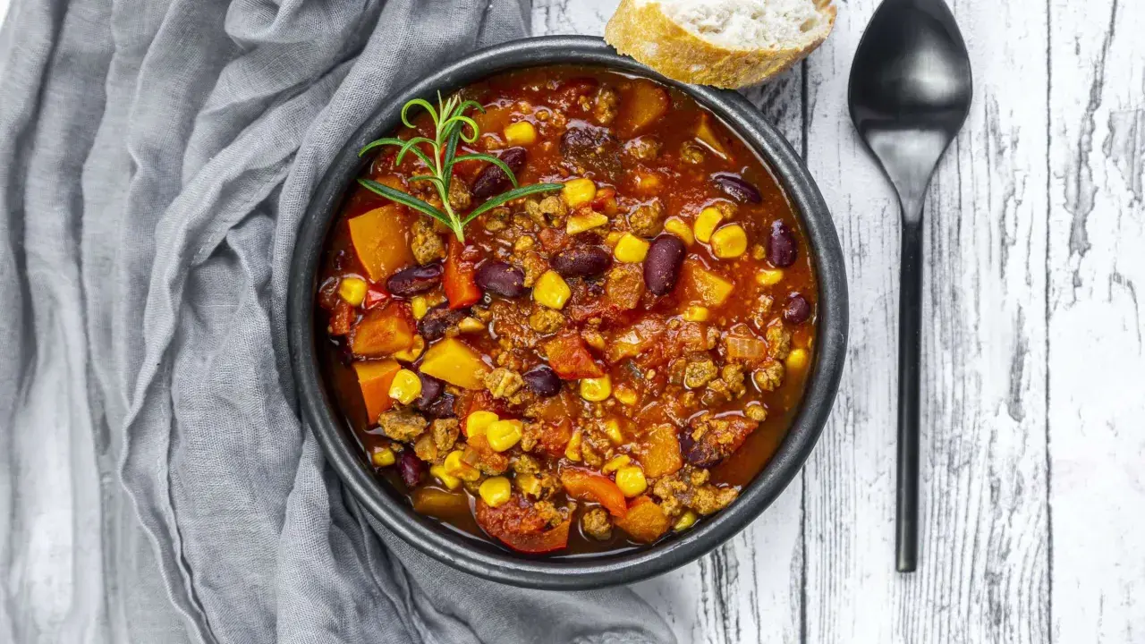 Herzhaftes Chili sin Carne mit Bohnen, Mais und Kürbis, garniert mit Rosmarin. Dazu ein Stück Brot und ein Löffel.