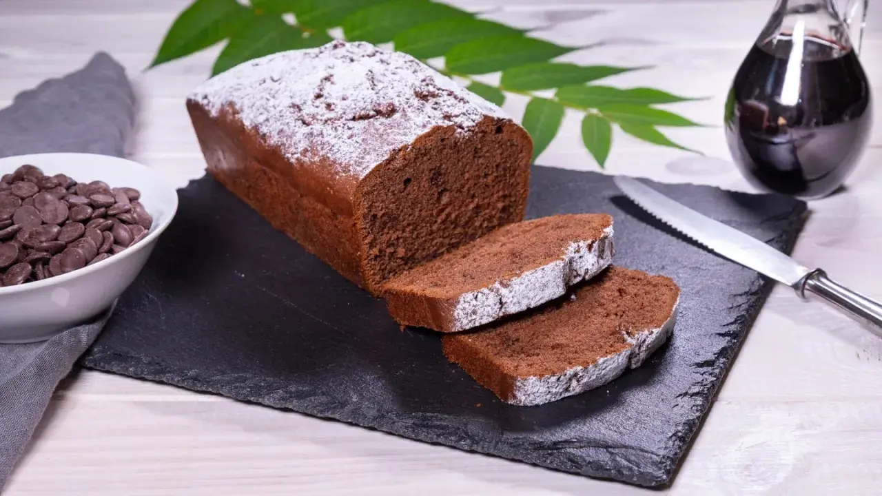 Ein saftiger Schokoladenkuchen, bestäubt mit Puderzucker, bereit für ein köstliches rotweinkuchen rezept. Daneben Schokoladendrops und ein Glas Rotwein.