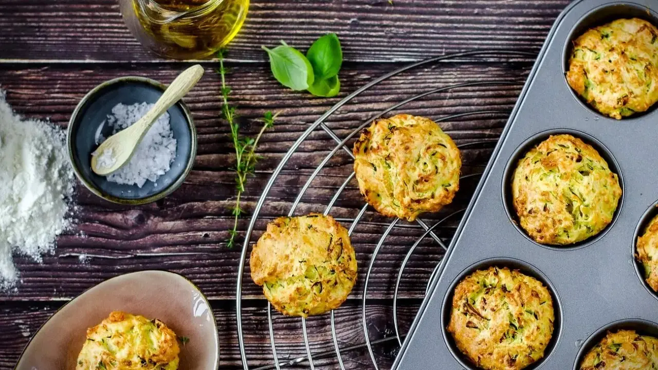 Goldbraune Zucchini-Muffins auf einem Gitter, einige in einer Muffinform. Zutaten wie Öl, Salz und Kräuter liegen bereit.