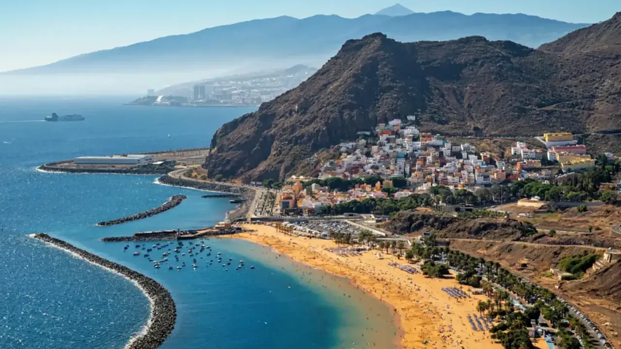 Bunte Häuser schmiegen sich an steile Hänge, während ein goldener Strand und das blaue Meer die Küste von Spanien zieren. Im Hintergrund erheben sich majestätische Berge.