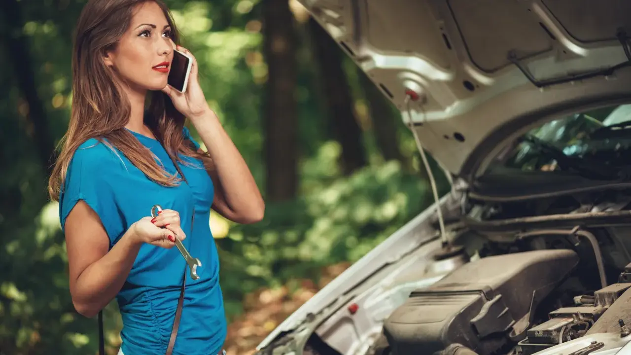 Mujer llama por teléfono con una llave inglesa en la mano, preguntando por qué se me para el coche en marcha. El capó del coche está abierto.