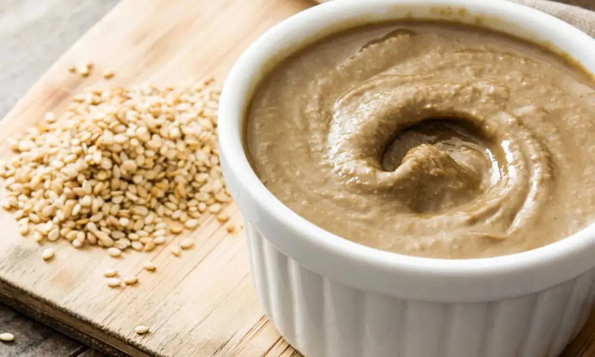 Un bol de sauce tahini crémeuse, à côté d'une pile de graines de sésame sur une planche en bois.
