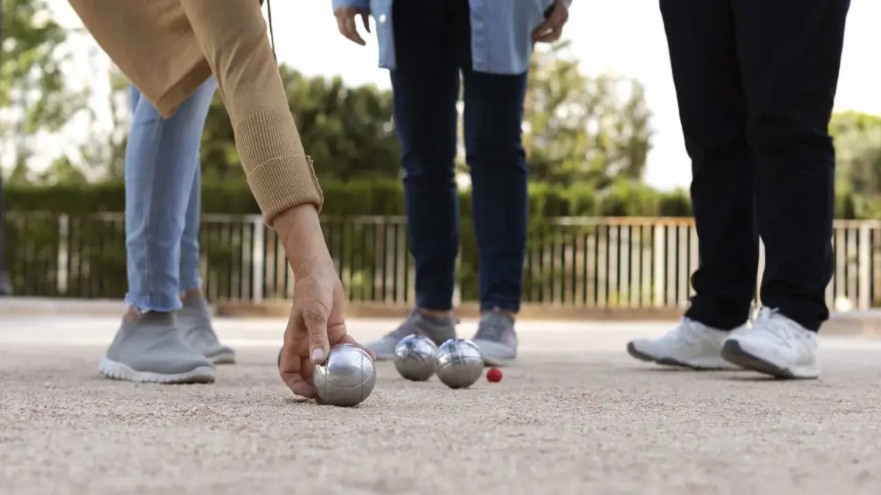 Une main de nom joueur de pétanque français saisit une boule argentée sur un terrain de gravier. D'autres boules et un cochonnet sont visibles.