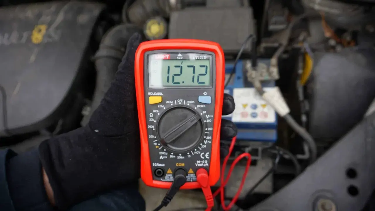 Ręka w czarnej rękawiczce trzyma multimetr pokazujący 12.72V, co pomaga sprawdzić akumulator.
