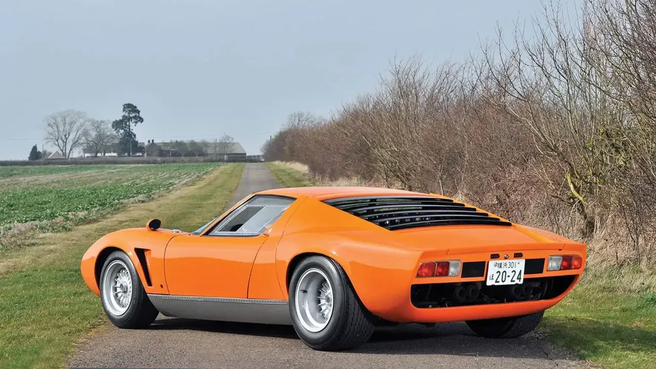 Ein leuchtend oranger Lamborghini Jota parkt auf einer einsamen Landstraße, umgeben von Feldern und kahlen Bäumen.