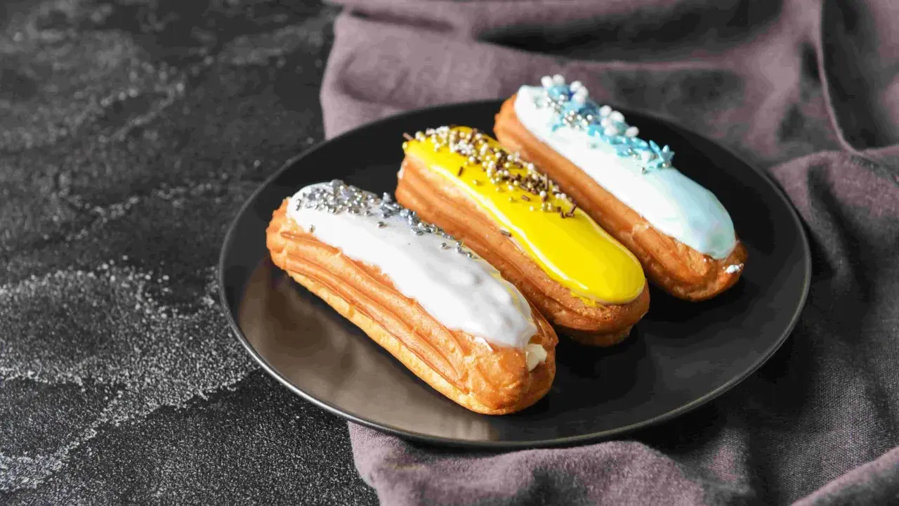 Trois éclairs gourmands sur une assiette noire : un blanc pailleté, un jaune croquant, un bleu ciel festif. La pâte à choux éclair inratable est prête à être dégustée.