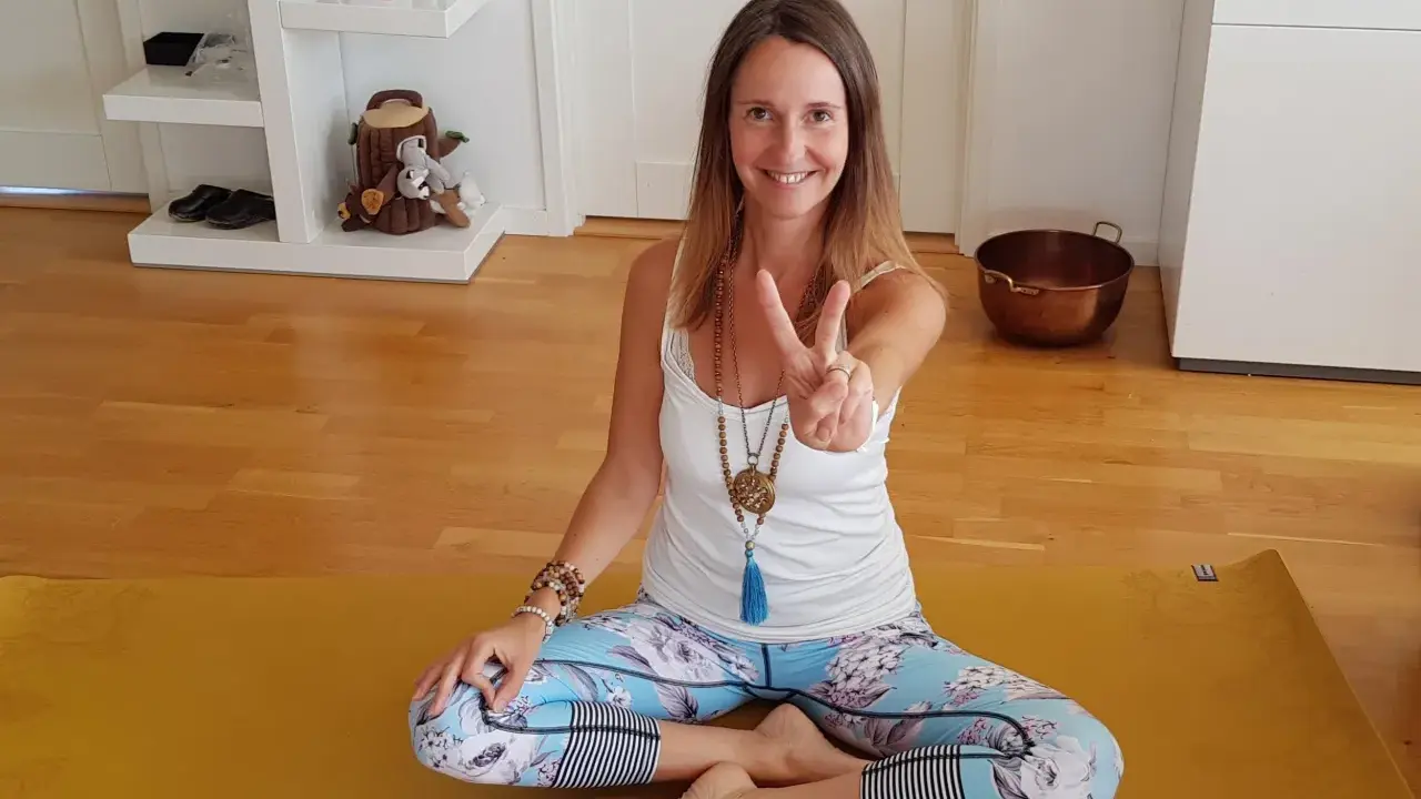 En kvinna sitter i lotusställning på en gul yogamatta, redo för morgon meditation. Hon ler och gör en fredstecken.