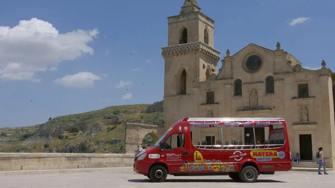 Un bus rosso "open top" per un tour di Matera, con la chiesa e i Sassi sullo sfondo.
