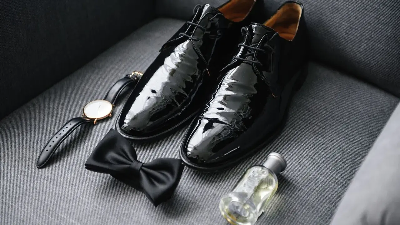 Shiny black patent leather shoes to wear with tuxedo, a bow tie, watch, and cologne are laid out on a grey couch.