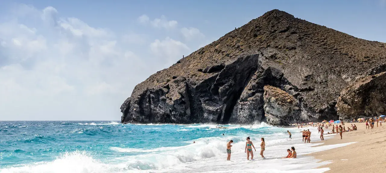 Sonniger Almería Urlaub: Menschen genießen den Strand und das Meer vor einer imposanten Felsformation.