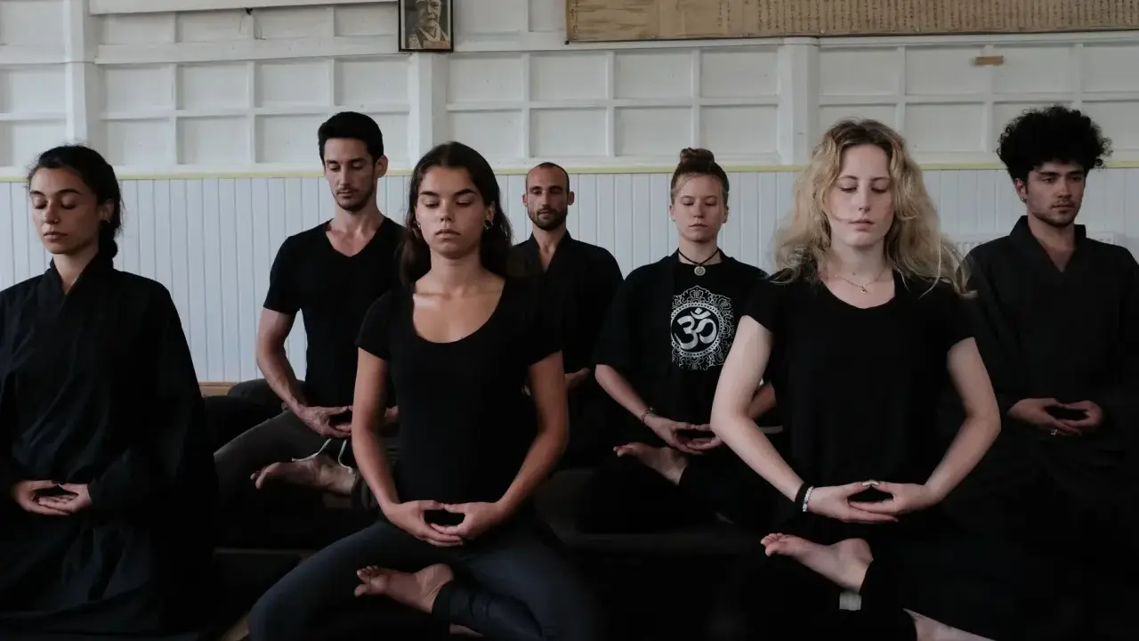 En grupp människor sitter i meditation, klädda i svarta kläder. En zen master leder sessionen.