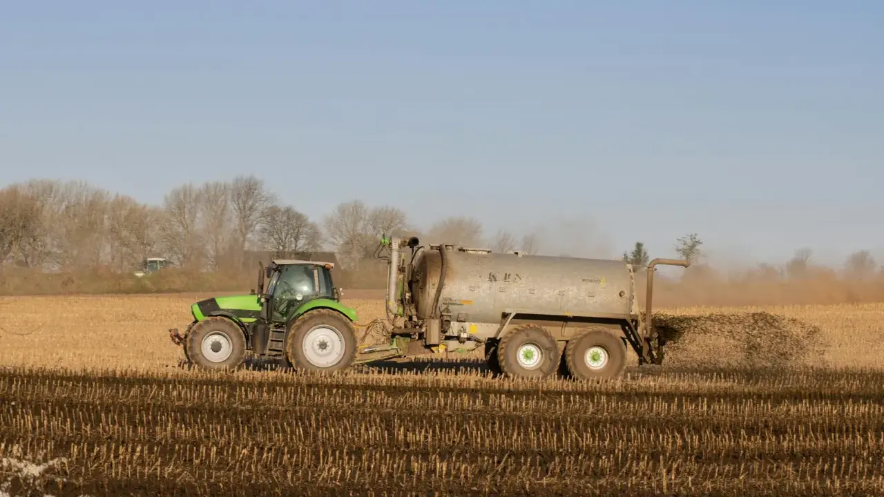 Zielony traktor z przyczepą rozprowadza obornik bydlęcy na polu.
